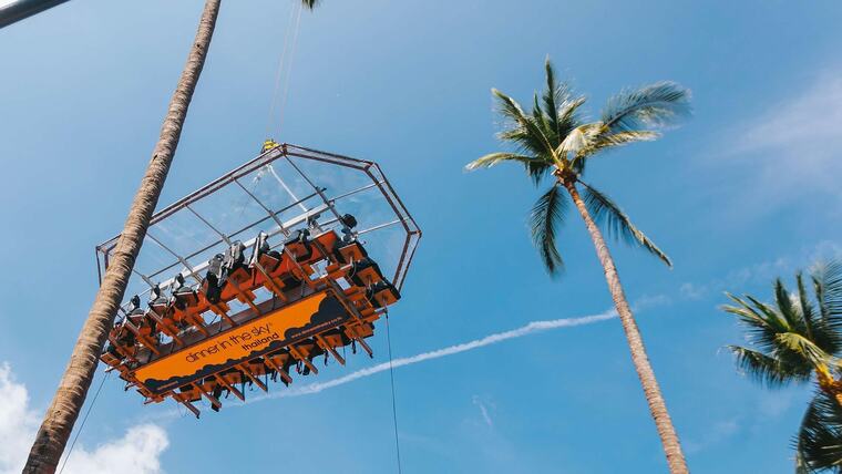 Стол Dinner in the Sky с гостями висит на тросах крана высоко над пальмами, вид снизу